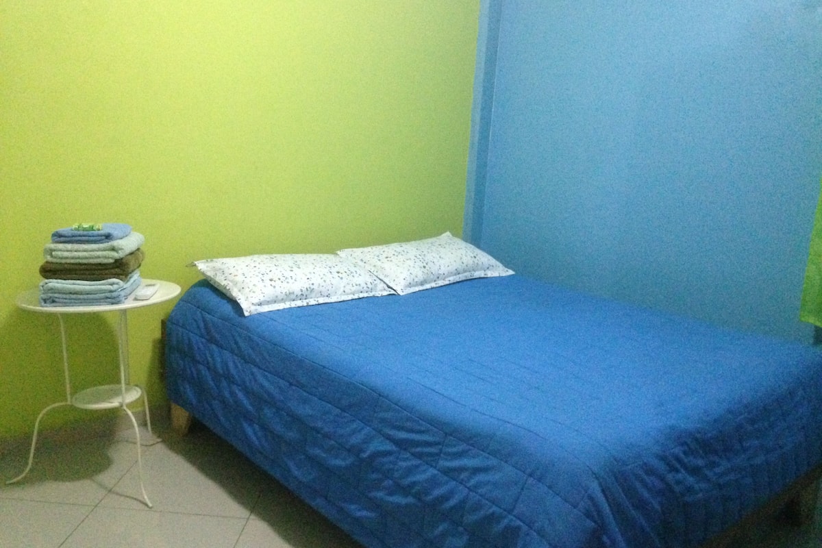 A small, single-bed room with a blue bedspread, a white pillow, and a stack of folded towels on a small white table.