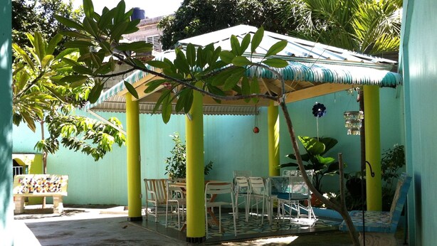 A covered outdoor seating area with a table and chairs, surrounded by greenery and a building with a blue and yellow facade. - Casa Azul Apartment (Puerto Plata)