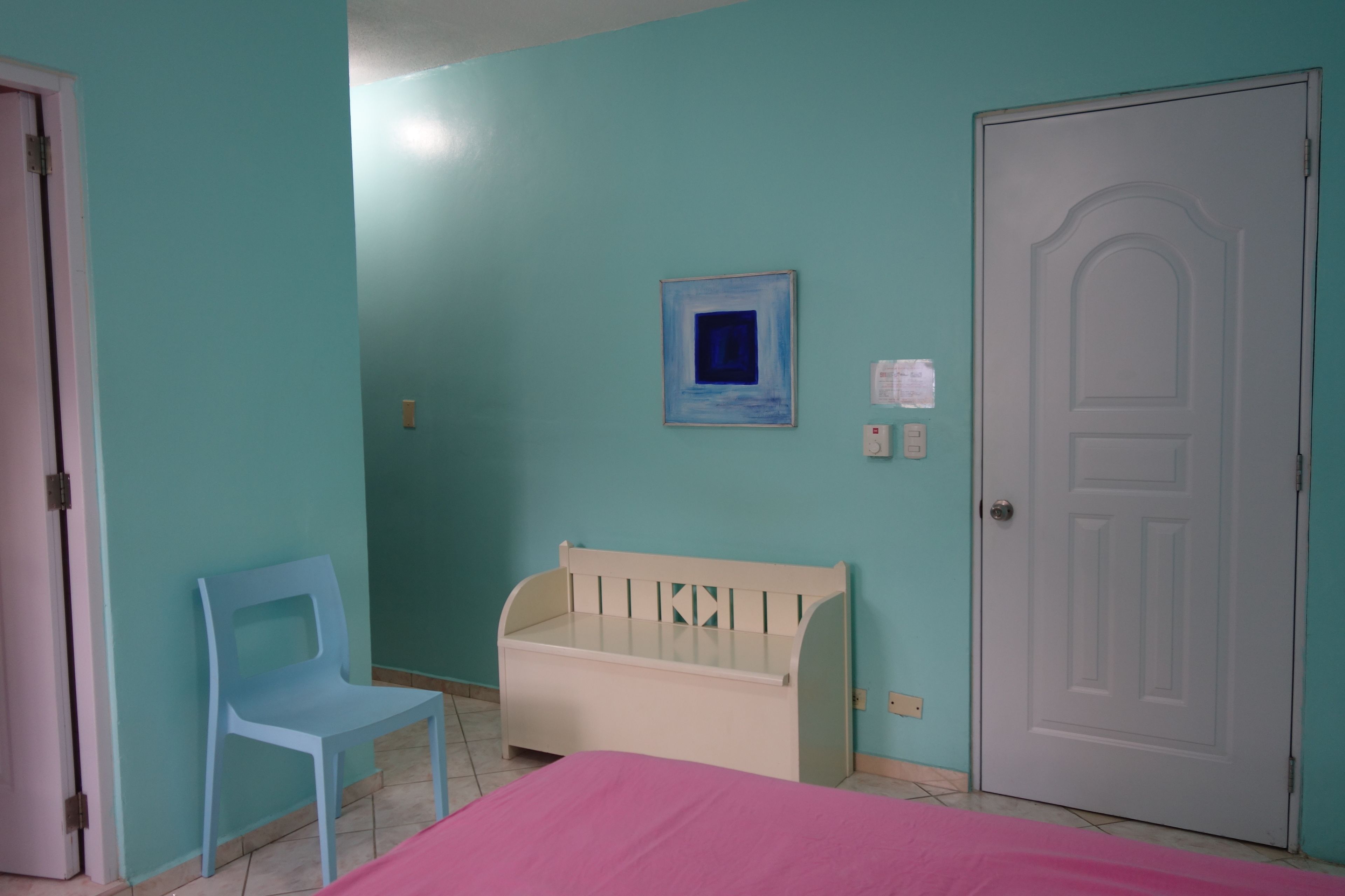 Ein Zimmer mit türkisfarbenen Wänden, einer weißen Tür, einem kleinen weißen Babybett, einem blauen Plastikstuhl und einem Bett mit rosa Bettwäsche.