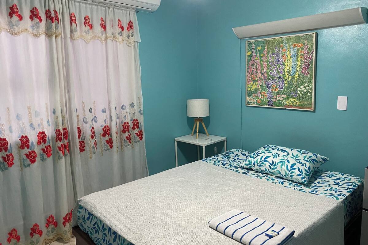 A bedroom with a bed, a nightstand, a lamp, a window with curtains, and a floral painting on the wall.