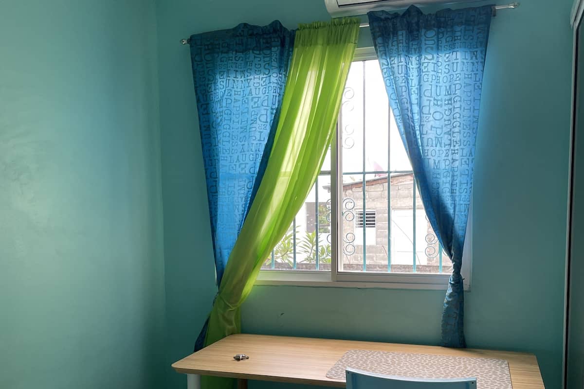 A small room with teal walls, a wooden desk, a blue chair, a bed with a white mattress, and a window with blue and green curtains.