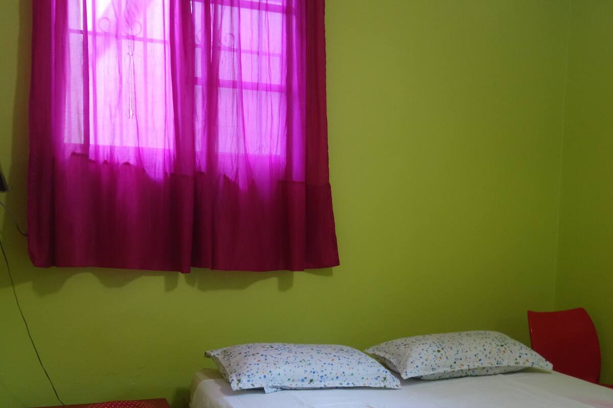A room with a bed, a nightstand, a red chair, and a window with pink curtains.