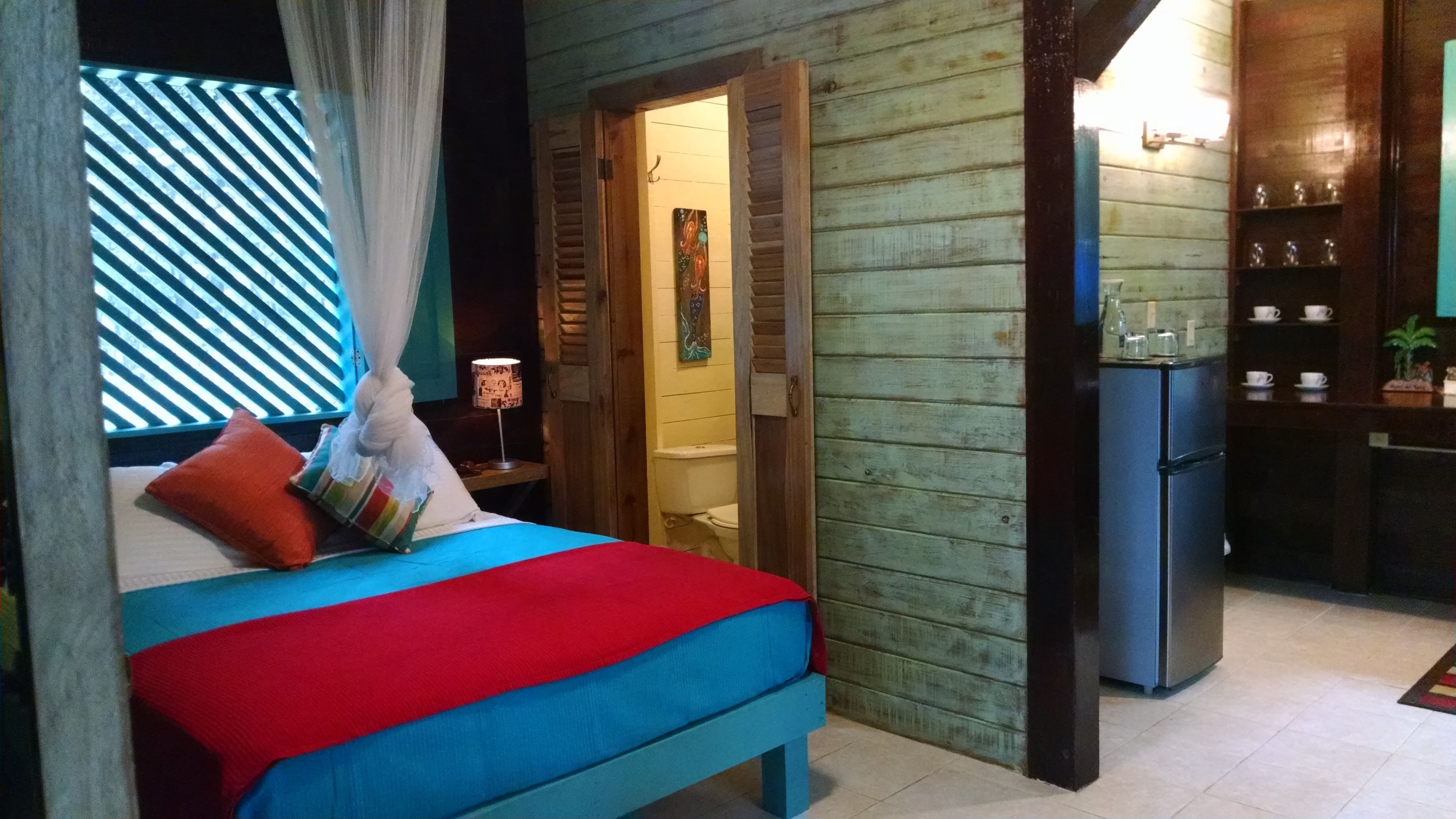 A room with a bed, a small table with a lamp, a refrigerator, and a shelf with cups and a plant.