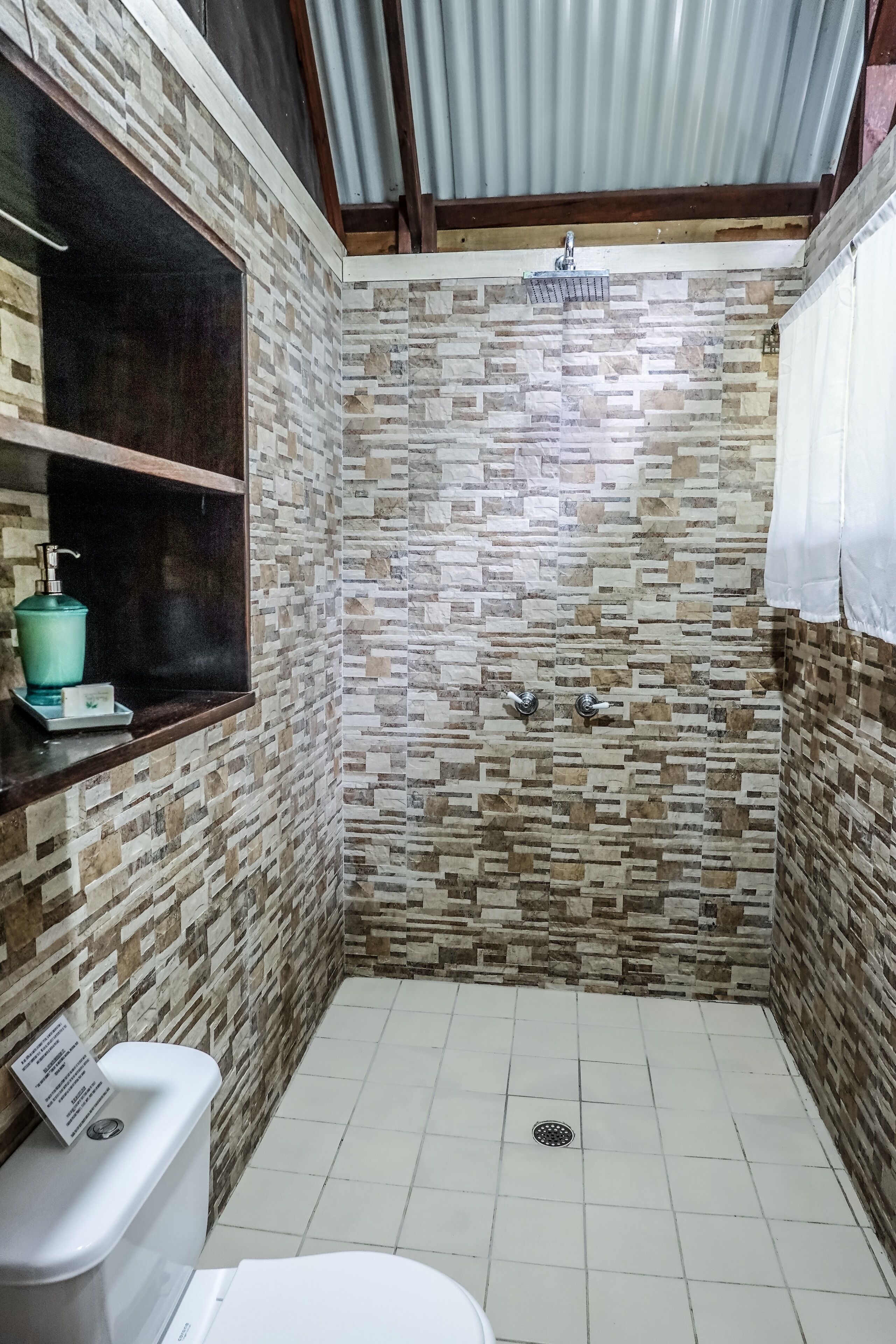 Casa de banho pequena, com azulejos, chuveiro, sanita e prateleira com artigos de higiene pessoal.