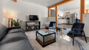 Un salon moderne avec un canapé, une table basse en verre et deux fauteuils. Un téléviseur est posé sur un meuble en bois, et une plante se trouve sur la gauche.