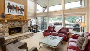 A spacious living area with a stone fireplace, leather furniture, and large windows offering a view of the outdoors. - Montaneros 416 - 4 Bedroom Platinum (Vail)