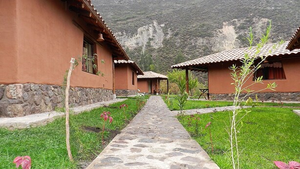 Un sendero conduce a una hilera de edificios de estilo tradicional con cimientos de piedra y techos de teja, rodeado de vegetación y un telón de fondo montañoso.