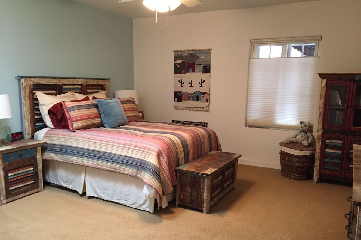 Een slaapkamer met een bed, een nachtkastje, een lamp, een raam met jaloezieën, een wanddecoratie en een klein houten kistje.