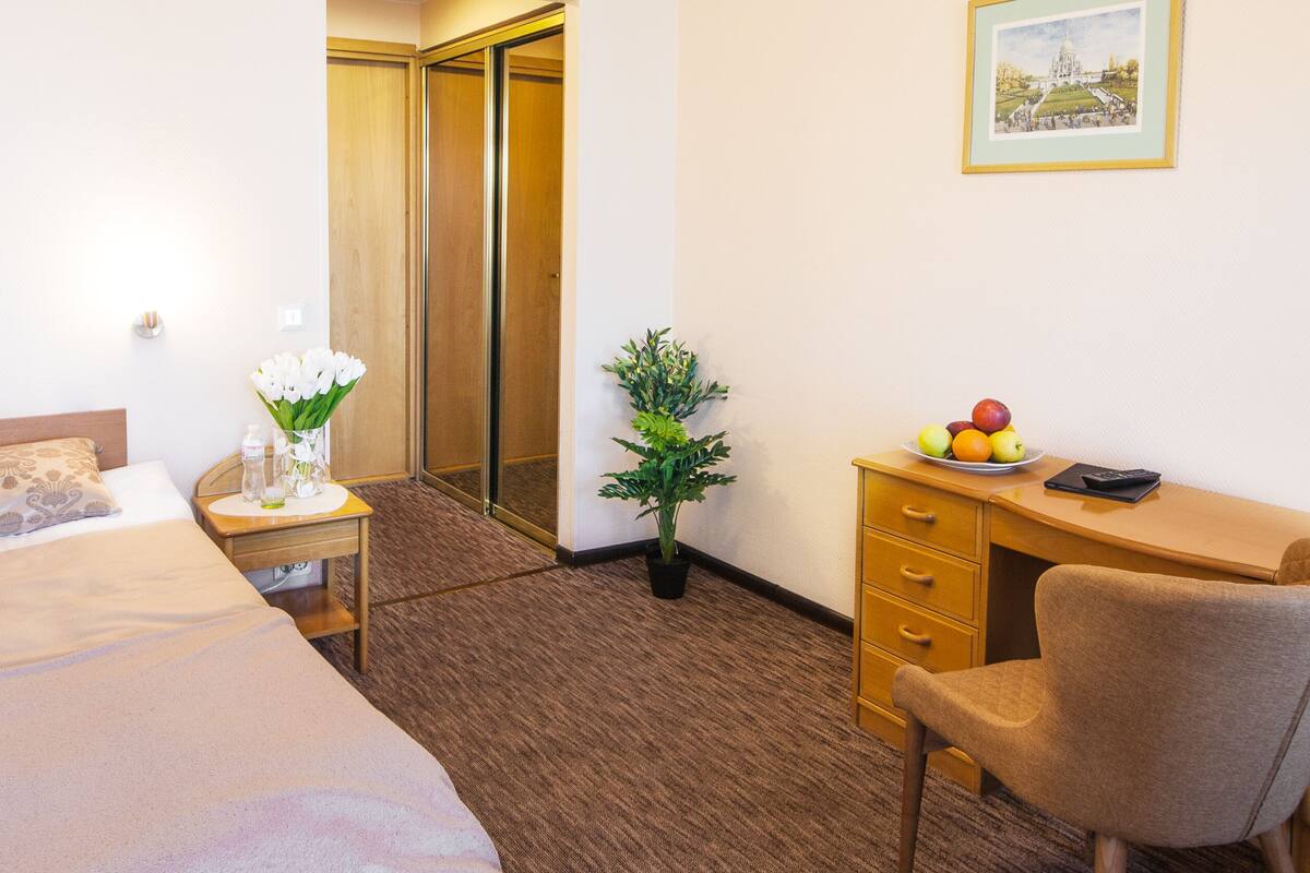 A hotel room with a bed, a desk, a chair, a small table with a vase of flowers, a fruit plate, and a framed picture on the wall.