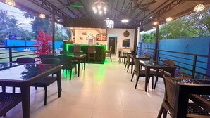 An outdoor cafe with tables and chairs, a counter with a green neon light, and a view of trees and a blue fence.