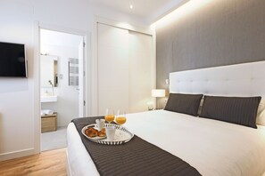 A hotel room with a large bed, a bedside table with a lamp, a tray with croissants and orange juice, and a TV mounted on the wall.