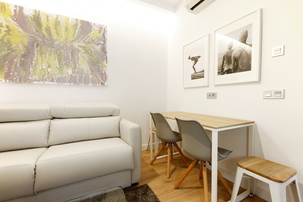A modern living room with a white leather sofa, a wooden dining table with chairs, and two framed pictures on the wall.