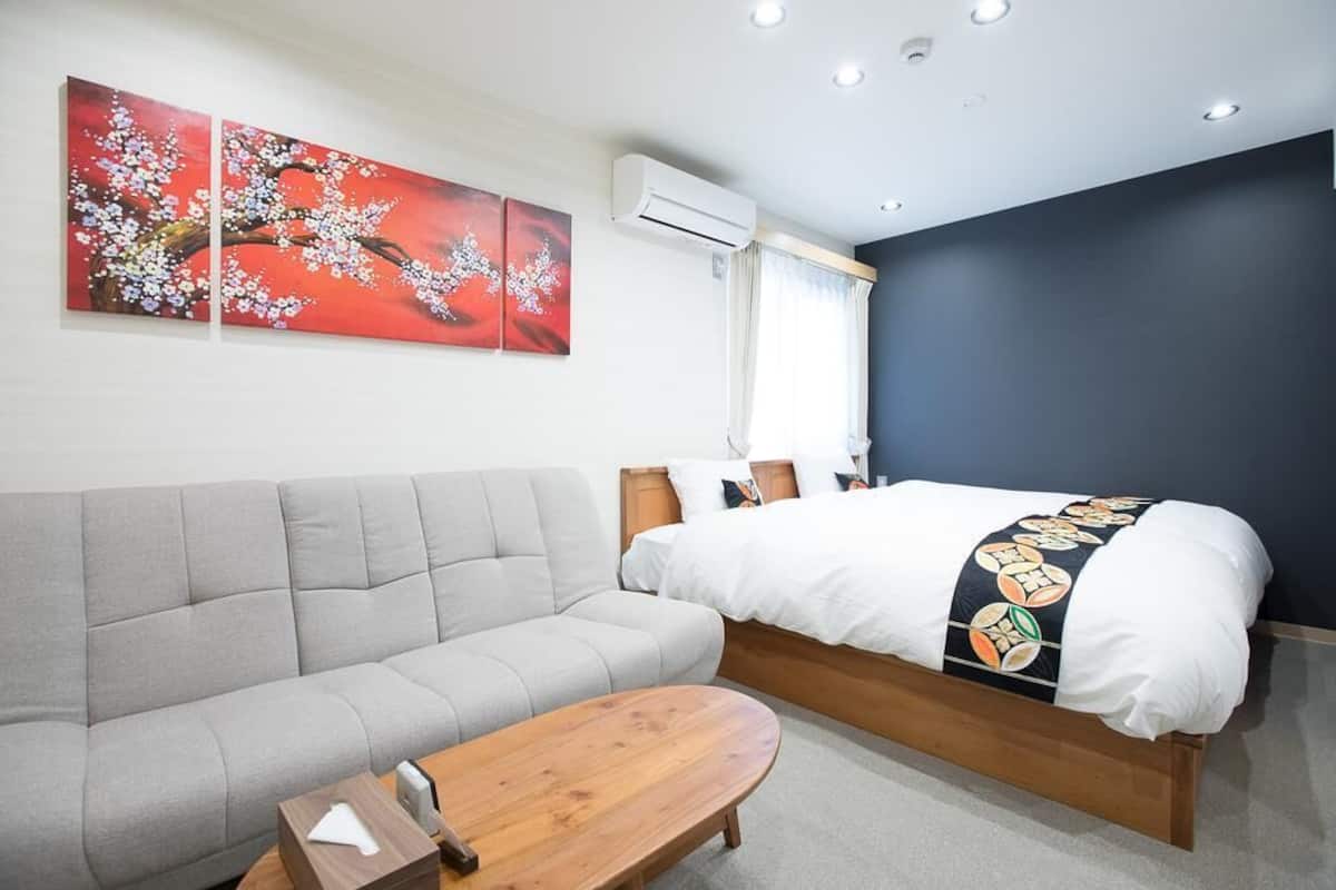 A hotel room with a bed, a sofa, a wooden side table, and two wall-mounted air conditioning units.