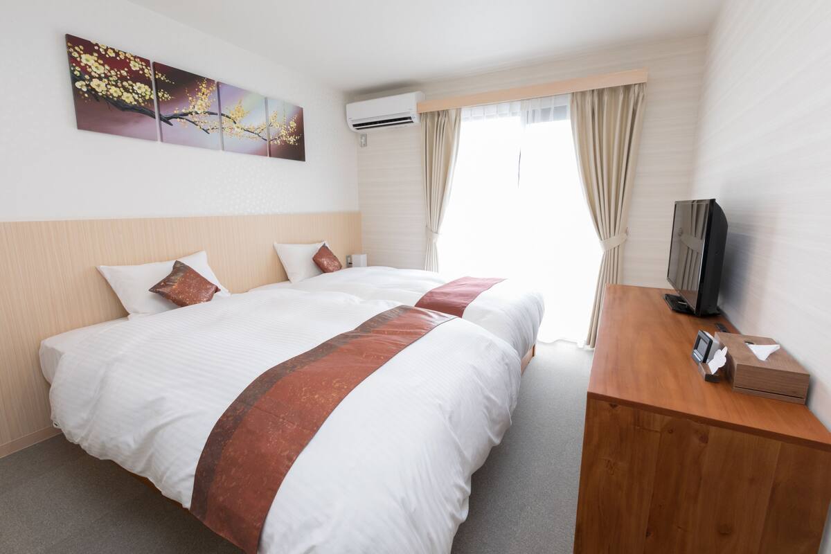 A hotel room with two beds, a wooden desk, a flat-screen TV, and a window with curtains.