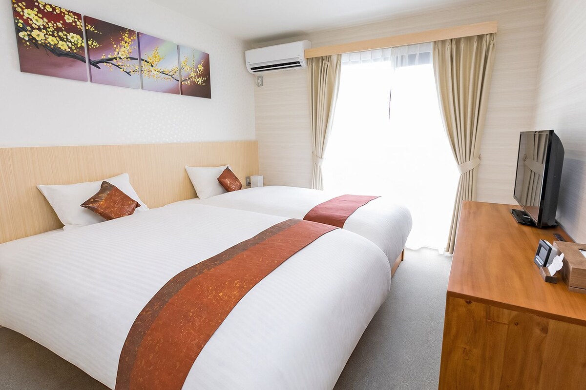 A hotel room with two beds, a wooden desk with a television, and a painting on the wall.