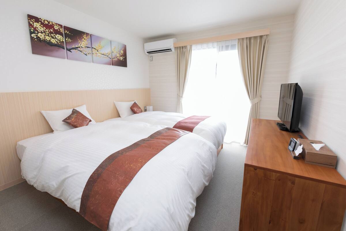 A hotel room with two beds, a wooden desk, a flat-screen TV, and a window with curtains.