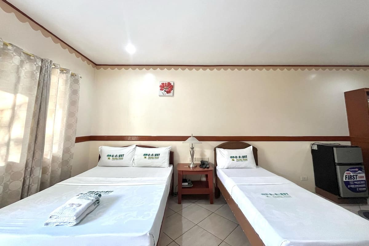 A hotel room with two single beds, a nightstand, a lamp, a refrigerator, and a wall-mounted fire alarm.