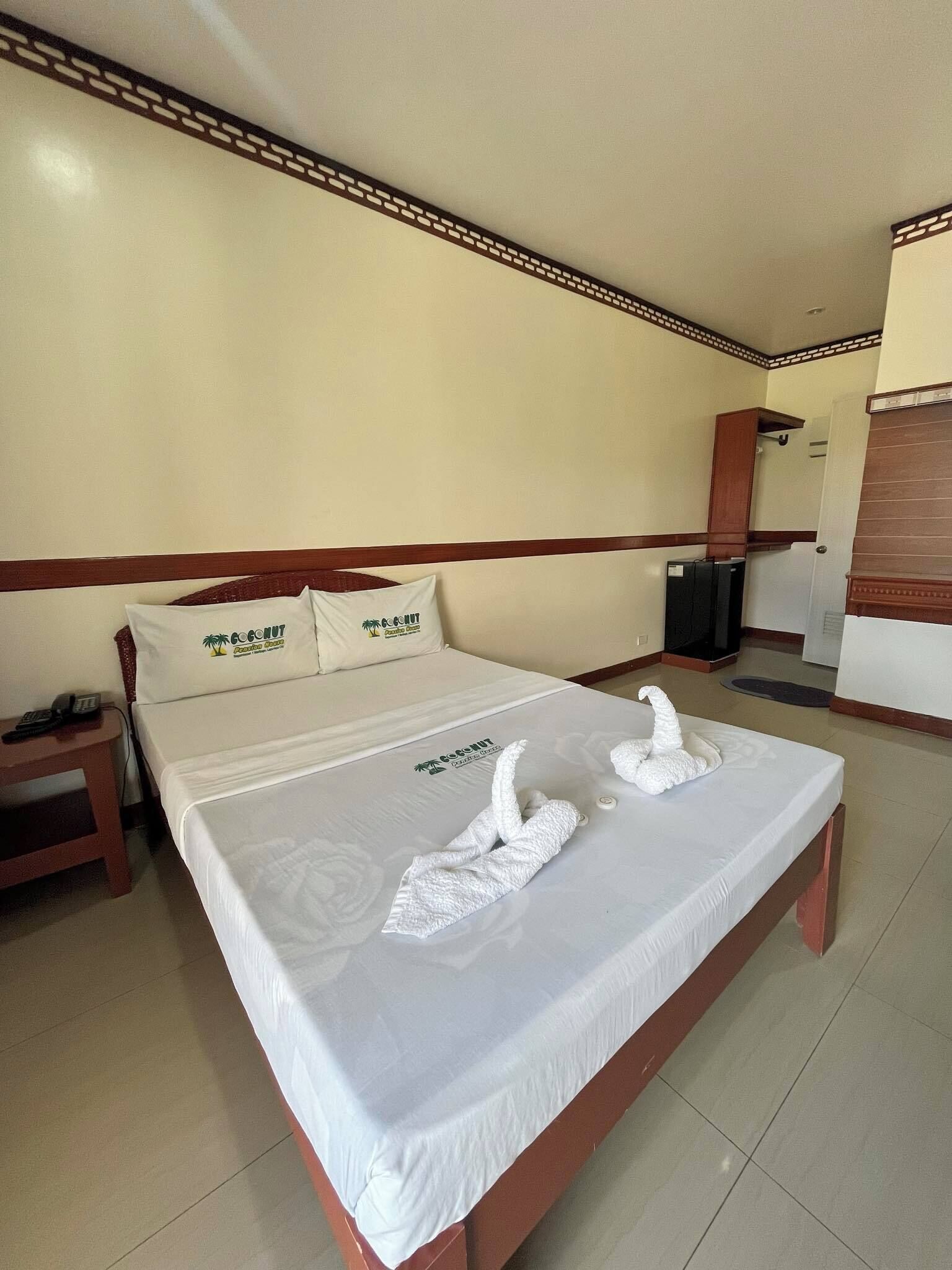 A hotel room with a neatly made bed, two pillows with a logo, a bedside table with a phone, and a small cabinet.