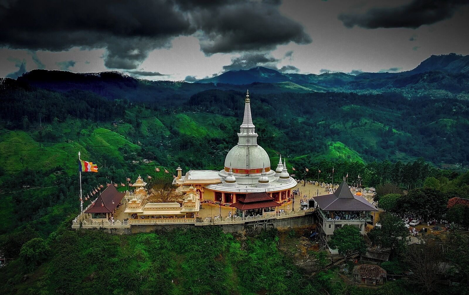 A large white temple complex surrounded by lush green hills, with a flagpole and a smaller building nearby.