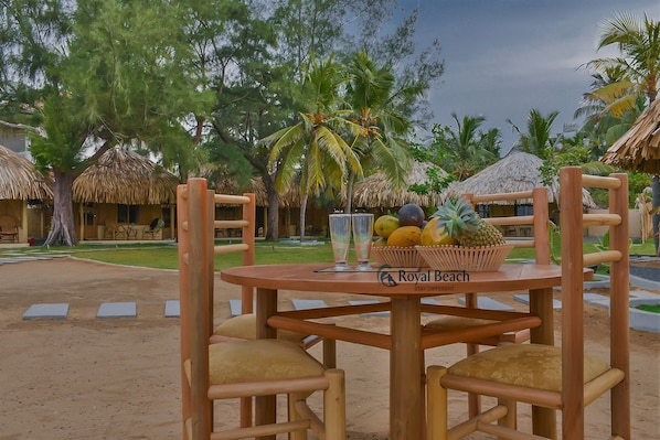 A wooden table with chairs and a fruit basket, surrounded by thatched-roof huts and palm trees.