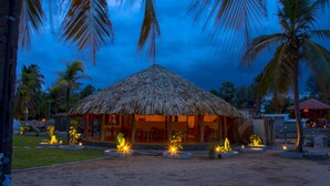A thatched-roof hut with a dining area and a poolside seating area, surrounded by palm trees and a sandy landscape. - Royal Beach Arugambay (Arugam Bay)