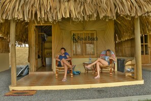 A covered outdoor seating area with two wicker chairs and a table, a sign reading "Royal Beach Stay Different", and a thatched roof structure.