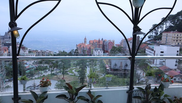 A balcony with potted plants and a view of a cityscape with buildings and a winding road.
