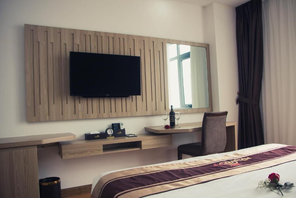Une chambre d’hôtel comprenant un lit, un bureau avec une chaise, un téléviseur fixé sur un panneau en bois, un miroir et une fenêtre avec des rideaux.