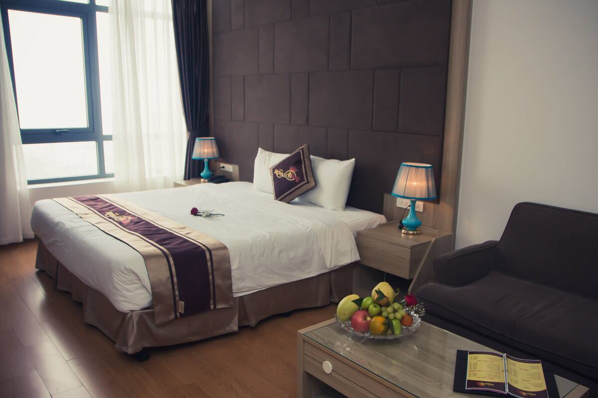 A hotel room with a large bed, a bedside table with a lamp, a coffee table with a fruit bowl, and a dark sofa.