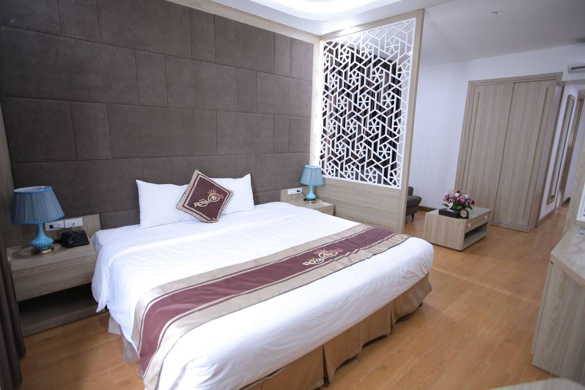 A neatly arranged bedroom with a large bed, two bedside tables with lamps, a wooden headboard, a decorative partition, and a wardrobe.