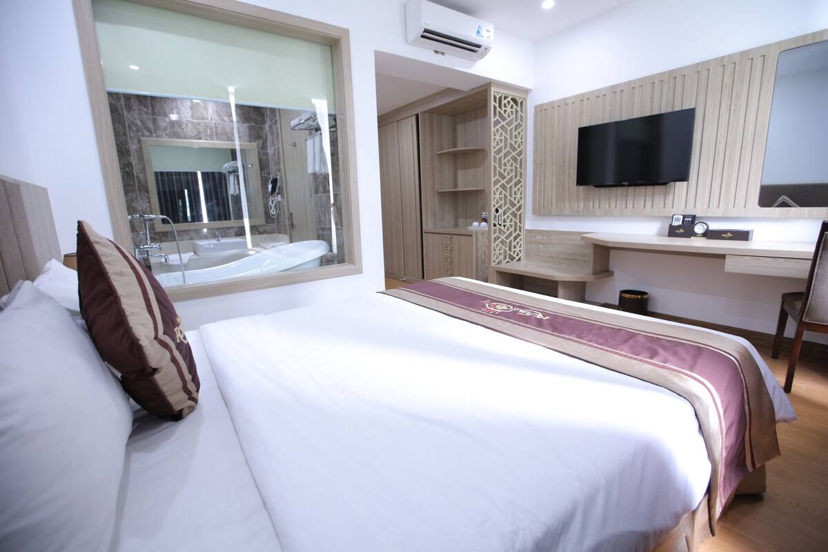 A hotel room with a bed, a desk, a TV, and a bathroom visible through a window.