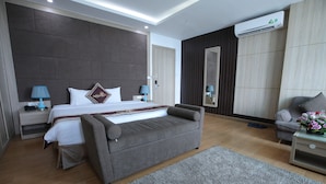 A modern hotel room with a large bed, a grey sofa, a small table with a lamp, and a large mirror.