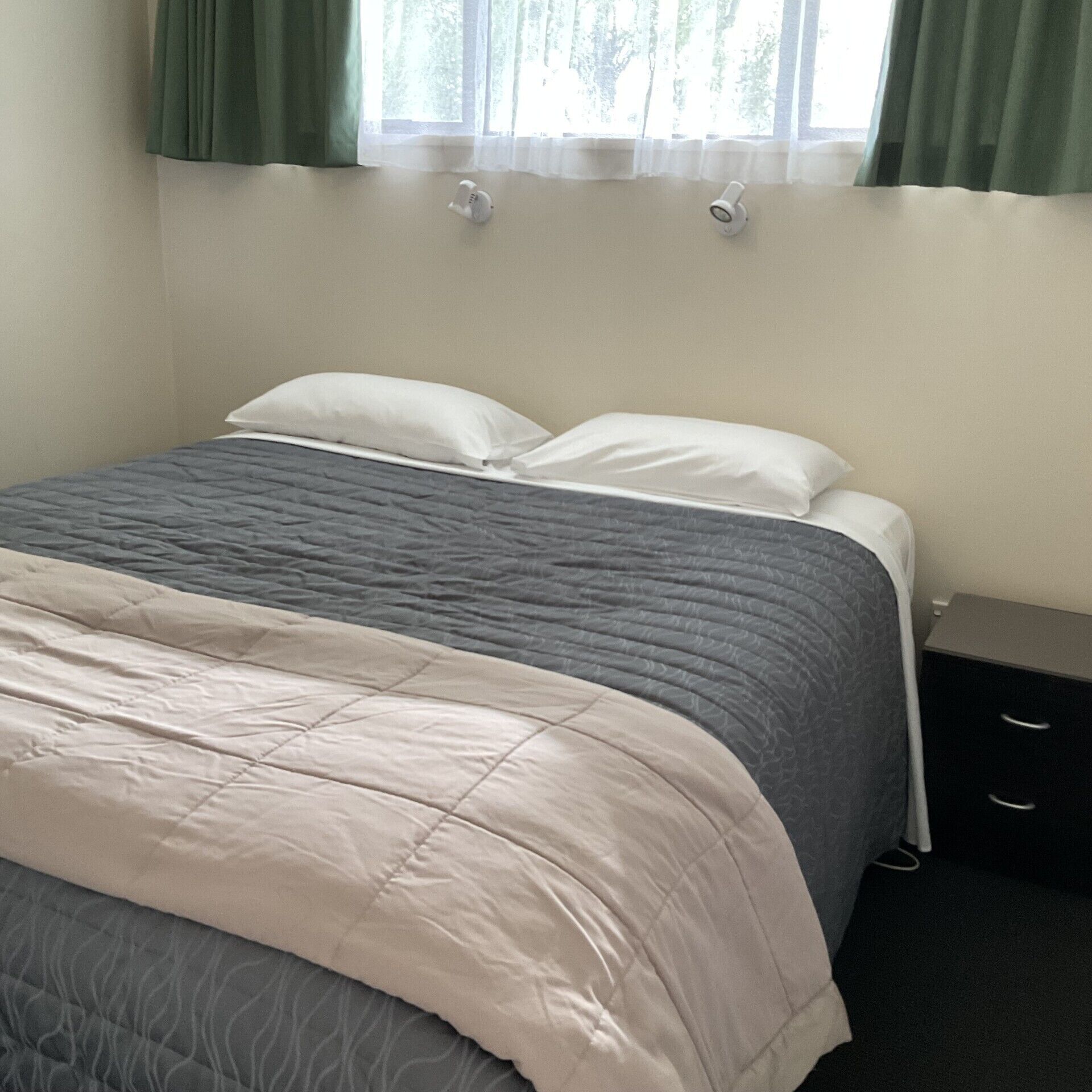 A neatly made bed with a grey and white duvet, a bedside table with drawers, and a window with green curtains.