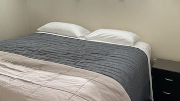 A neatly made bed with a grey and white duvet, a bedside table with drawers, and a window with green curtains.