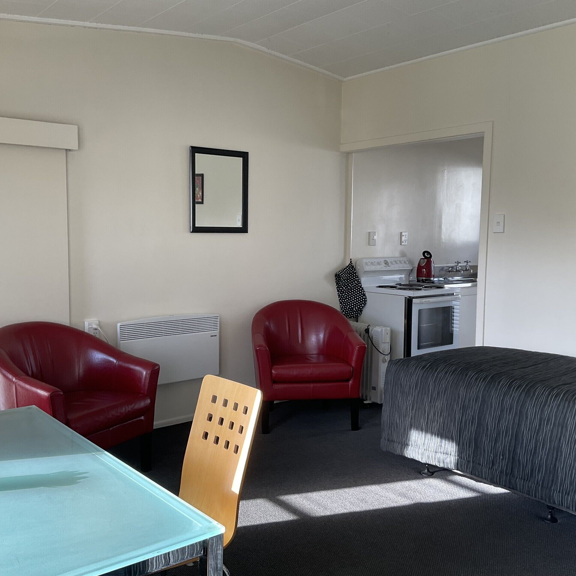 Ein kleines Hotelzimmer mit einem Bett, zwei roten Ledersesseln, einem kleinen Tisch, einer Küchenzeile und einem Spiegel an der Wand.