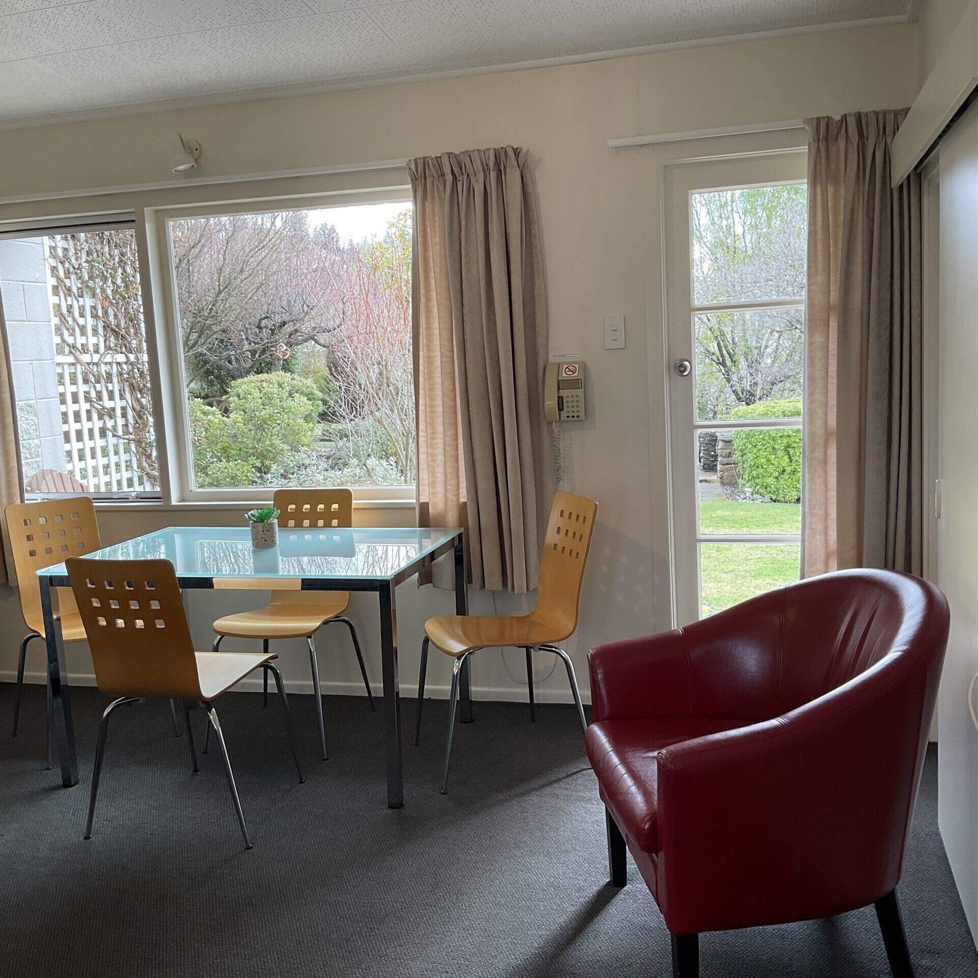 A room with a glass table, four chairs, a red chair, and large windows with curtains.