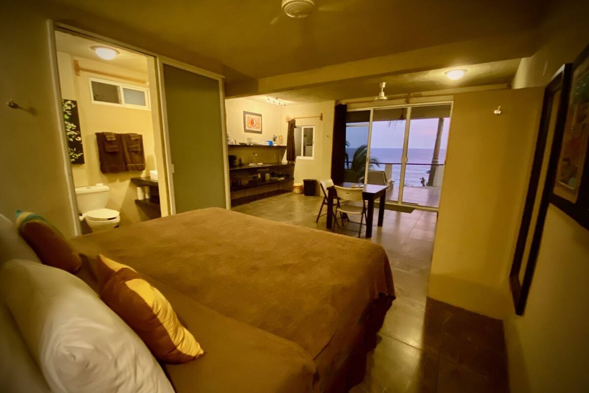 A bedroom with a large bed, a bathroom, and a balcony with a view of the ocean.