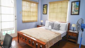 A bedroom with a wooden bed, two bedside tables, a chair, a standing fan, and two windows with blinds.