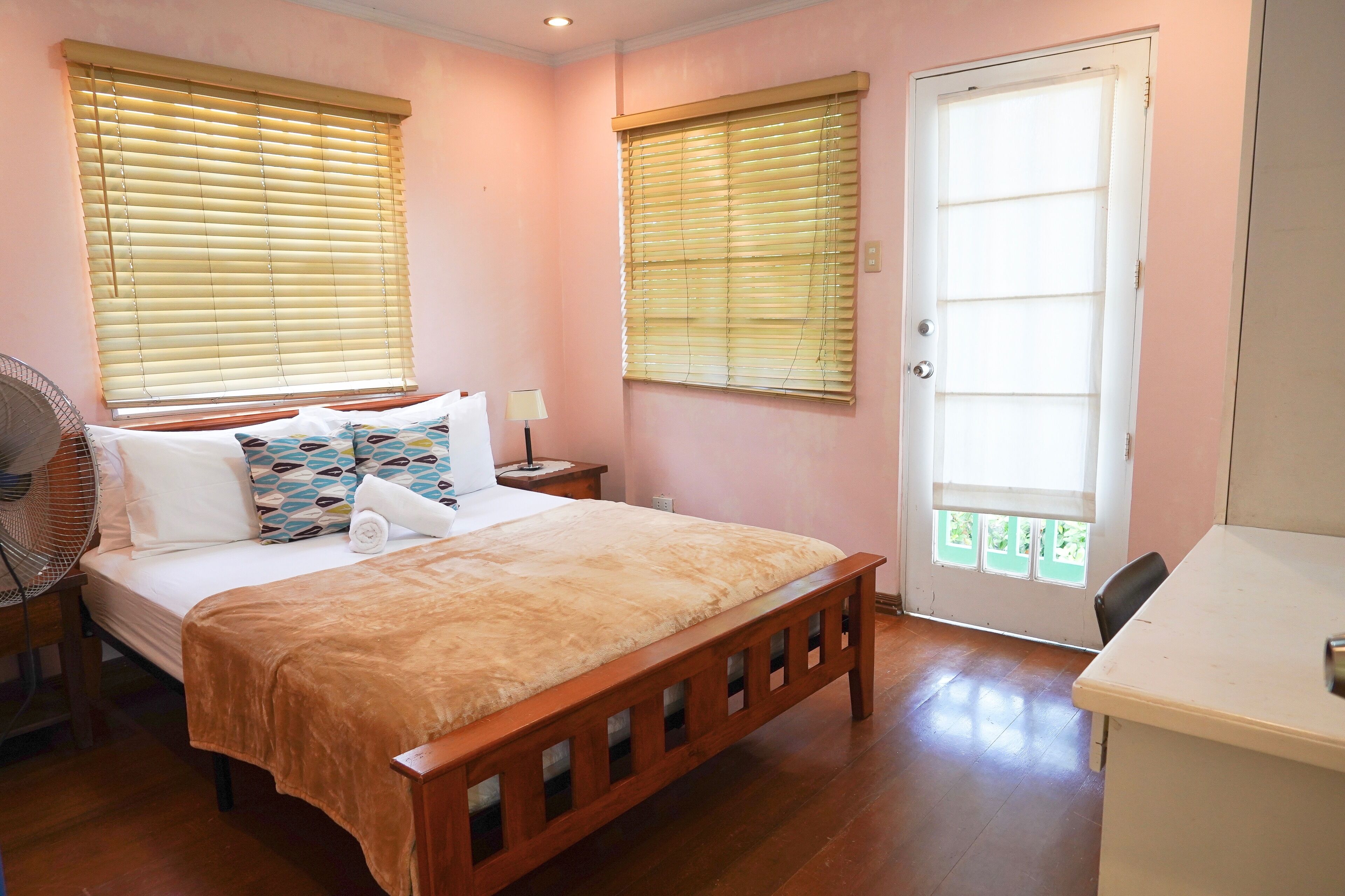 Un dormitorio con una cama de madera, dos mesitas de noche, una mesita auxiliar, un ventilador, un escritorio y una puerta con vistas.