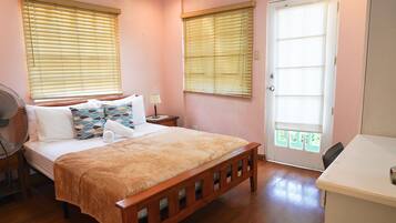 Un dormitorio con una cama de madera, dos mesitas de noche, una mesita auxiliar, un ventilador, un escritorio y una puerta con vistas.