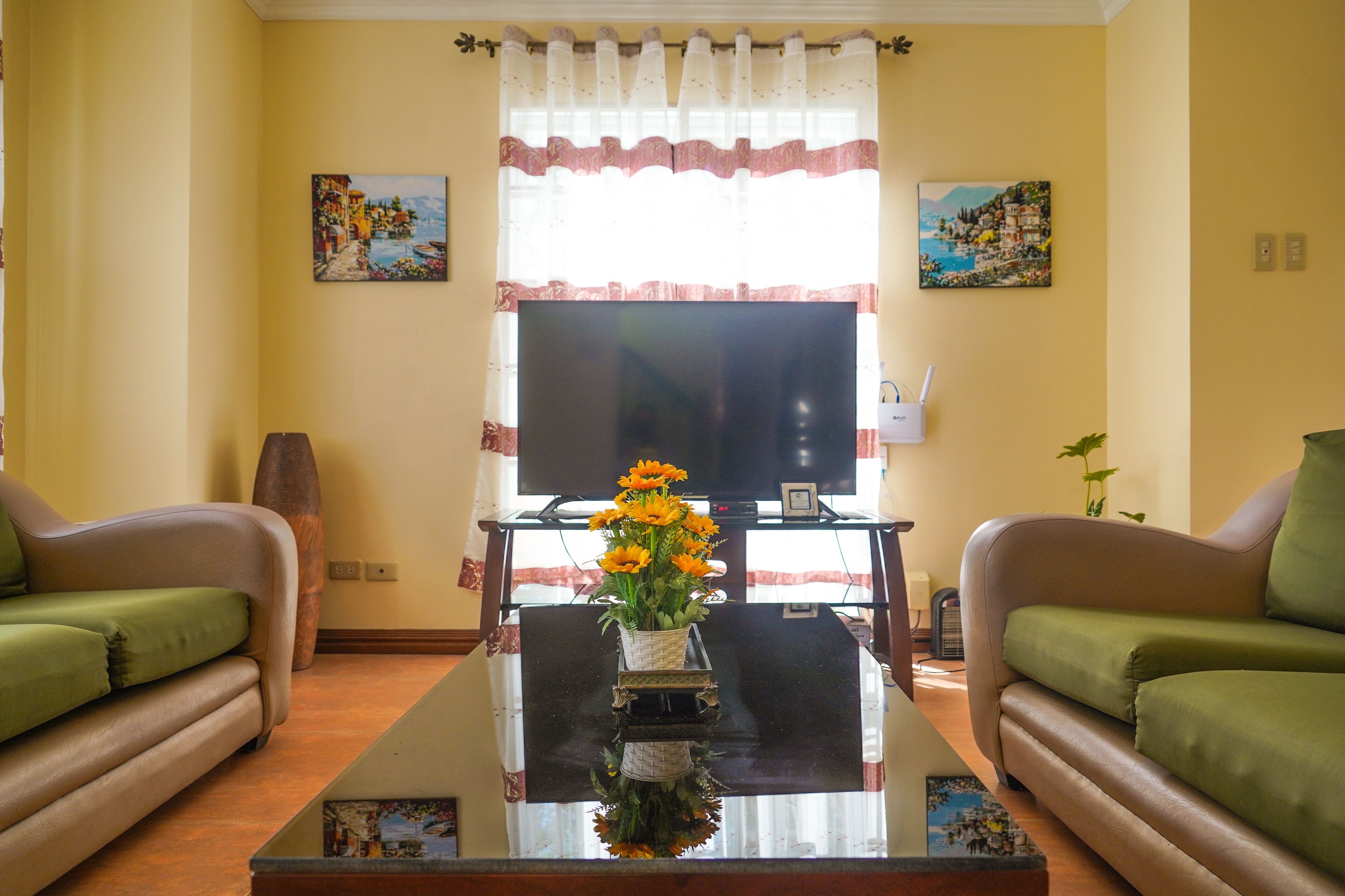 Ein Wohnzimmer mit einer Couch, einem Couchtisch mit Blumenvase, einem Flachbildfernseher und zwei Bildern an der Wand.