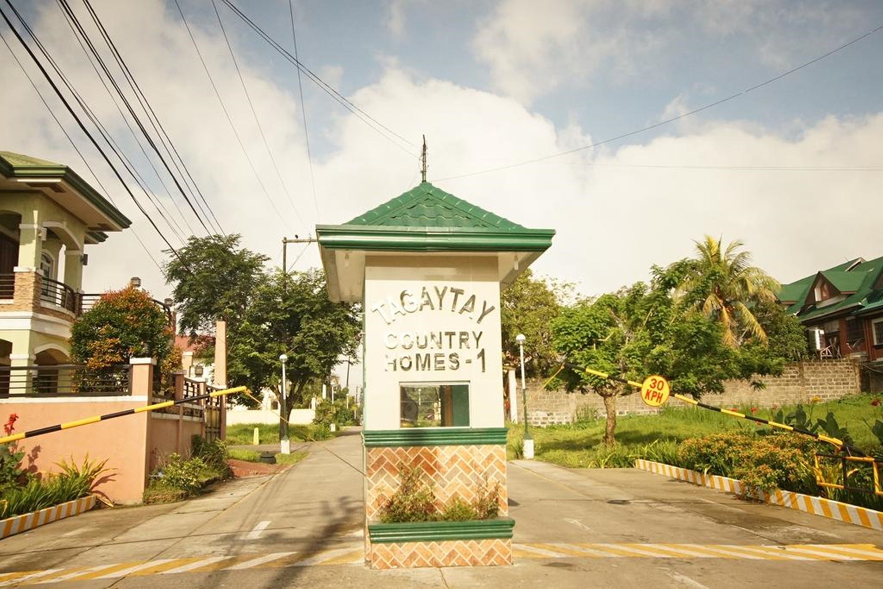 A gated entrance with a sign for Tagaytay Country Homes-1, surrounded by greenery and residential buildings.