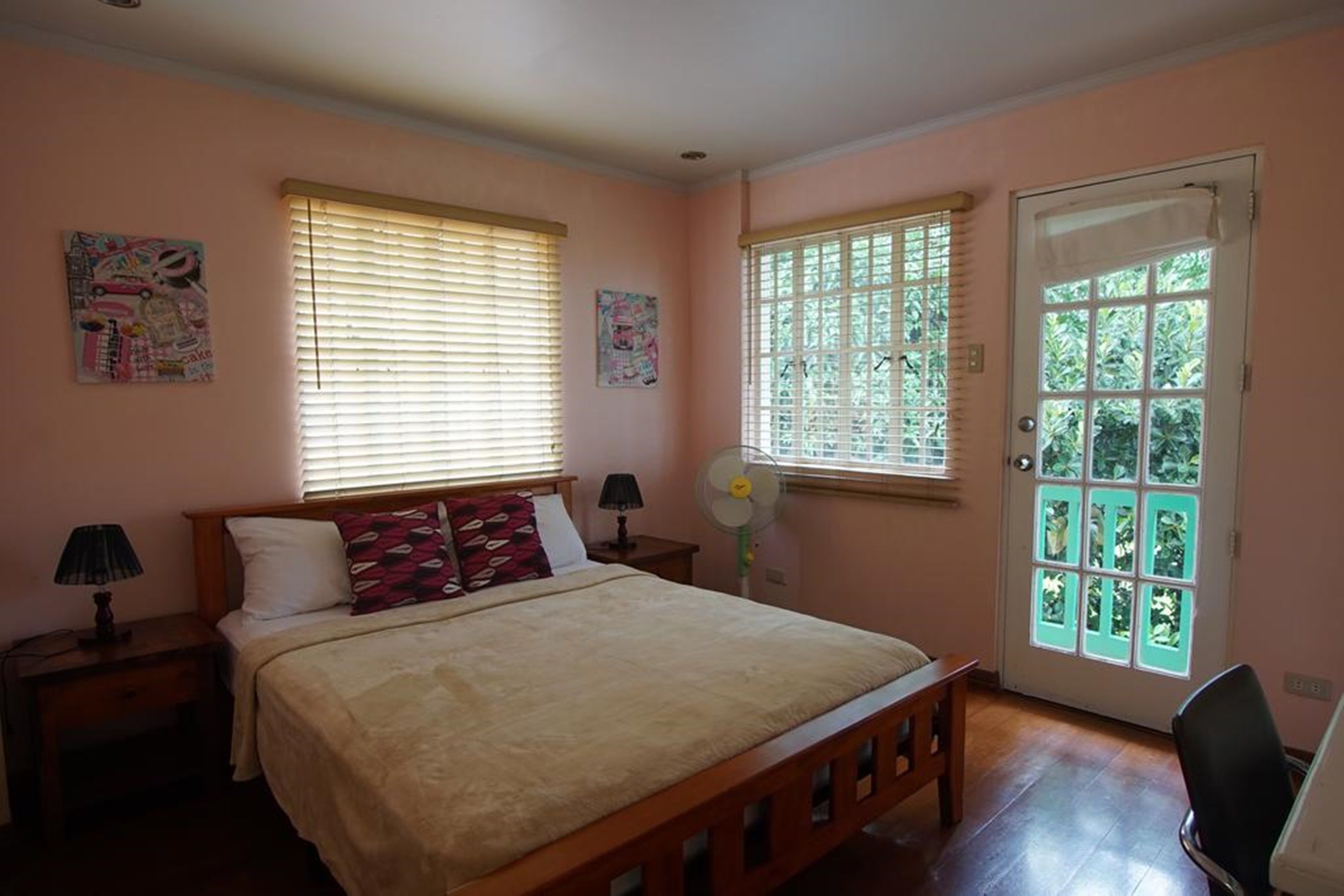 A bedroom with a bed, two bedside tables with lamps, a window with blinds, a door with a view, and wall art.