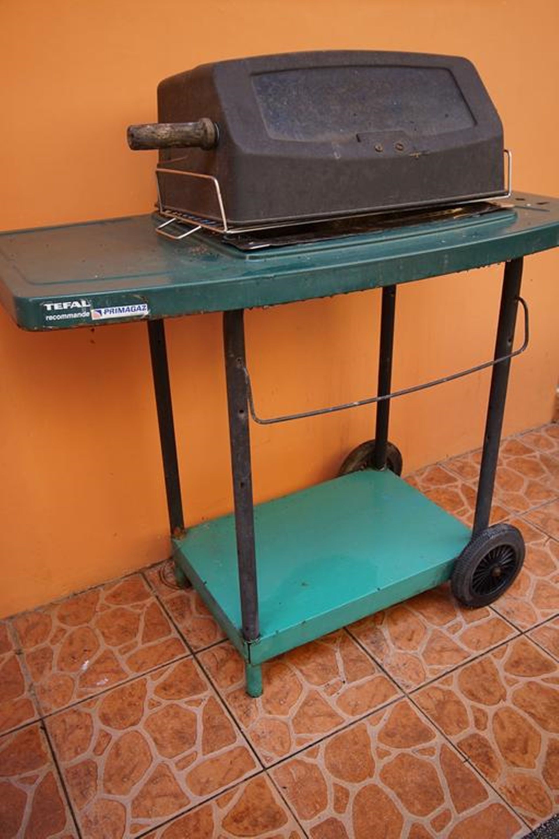 A portable grill on a green cart with wheels, placed on a tiled floor against an orange wall.