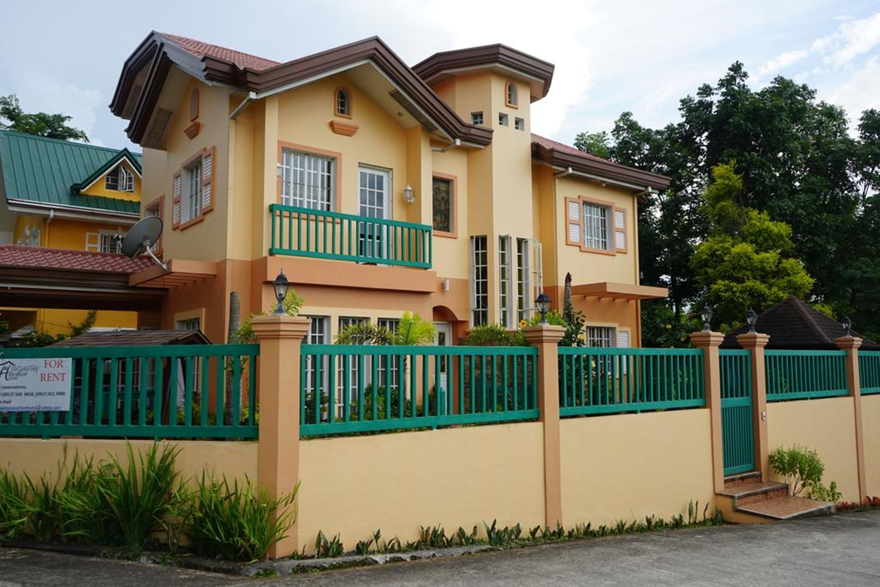 A two-story house with a green fence, a balcony, and a sign indicating it is for rent.