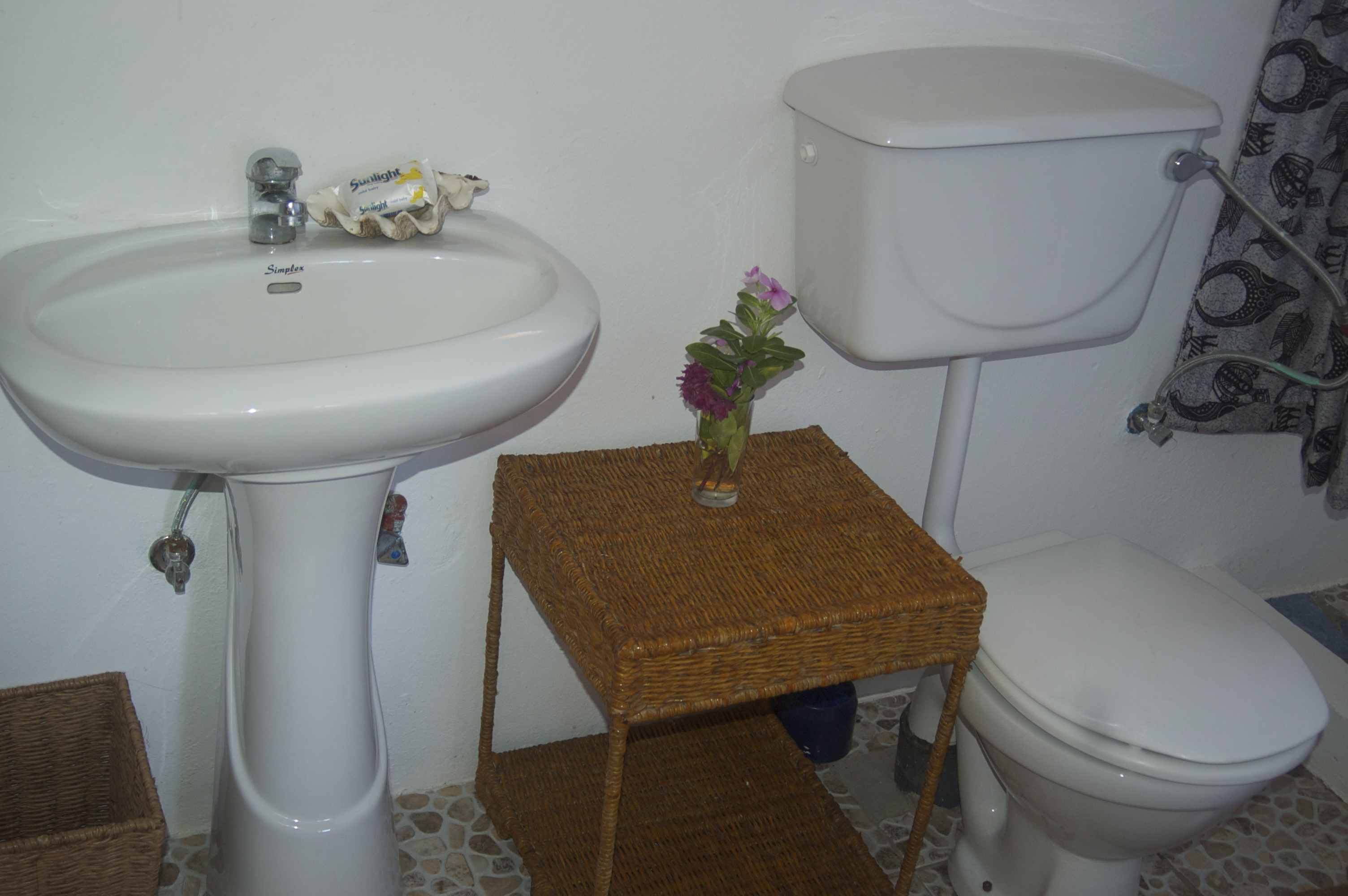 Un baño con un lavamanos blanco, inodoro y un pequeño taburete de mimbre con un jarrón de flores.