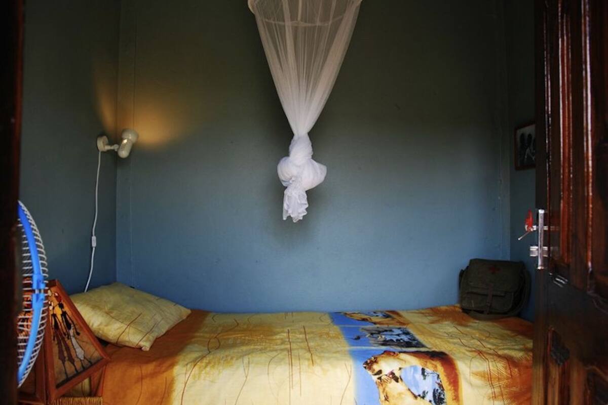 Una habitación con una cama, mosquitera, una silla y una lámpara fijada a la pared.