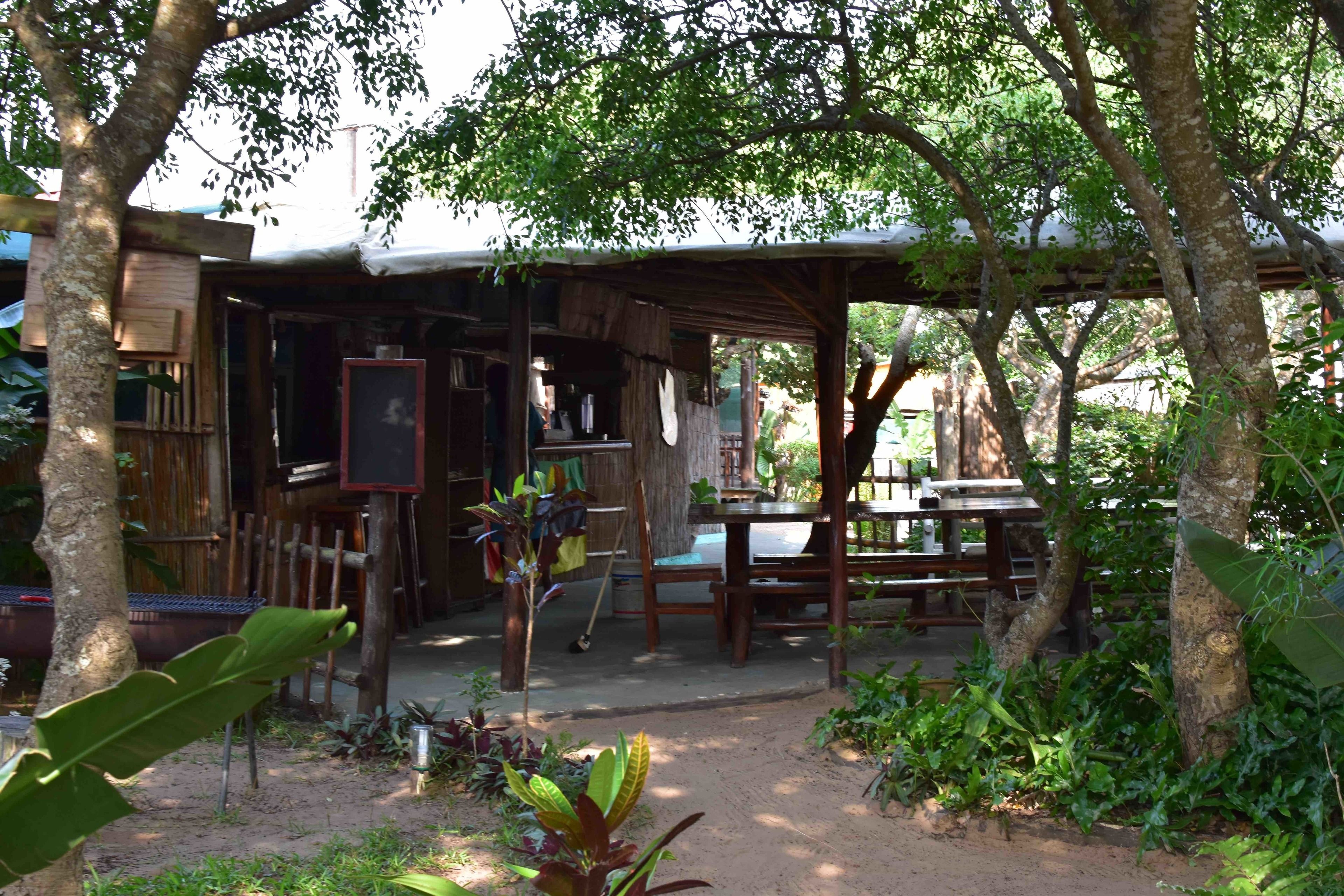A rustic wooden structure with a covered seating area and a small table, surrounded by trees and plants.