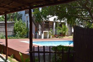 A covered outdoor area with a swimming pool, surrounded by plants and a wooden fence. - Gamboozini Lodge (Ponta d'Ouro)