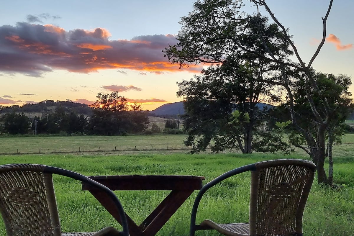 Vista de uma mesa com duas cadeiras de vime voltada para um campo gramado, árvores e um pôr do sol ao fundo.